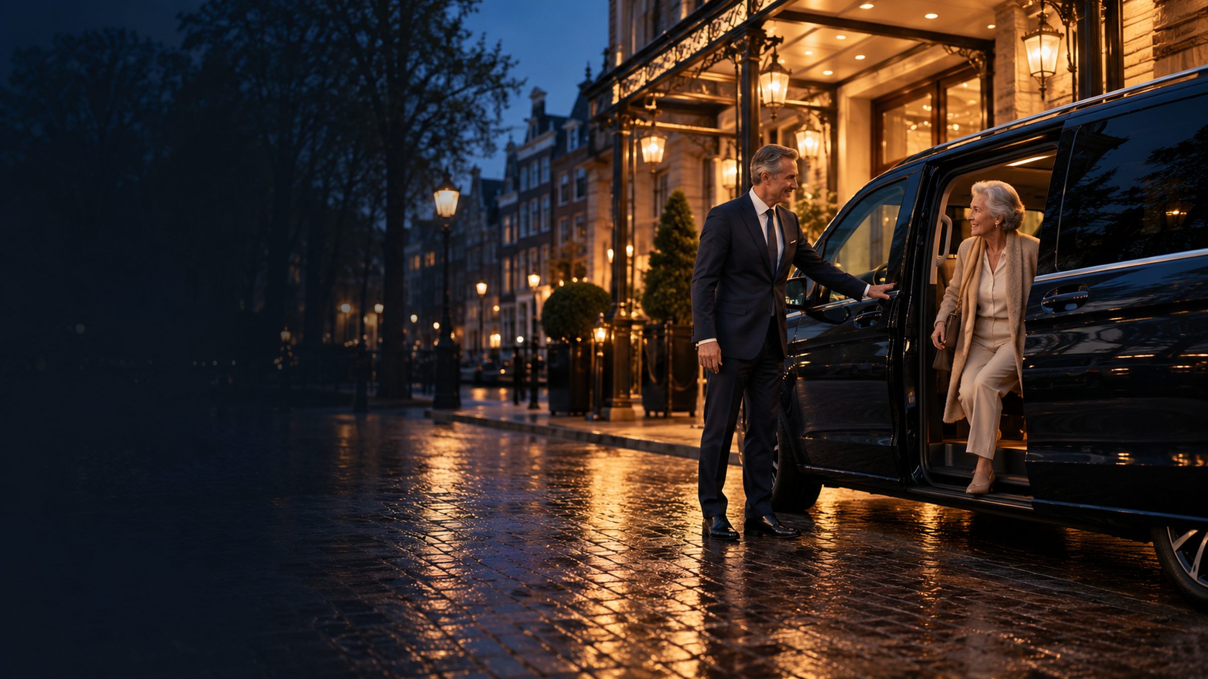 Een chauffeur houdt de deur open van een donkere, rolstoeltoegankelijke personenbus voor de ingang van een hotel bij schemering.